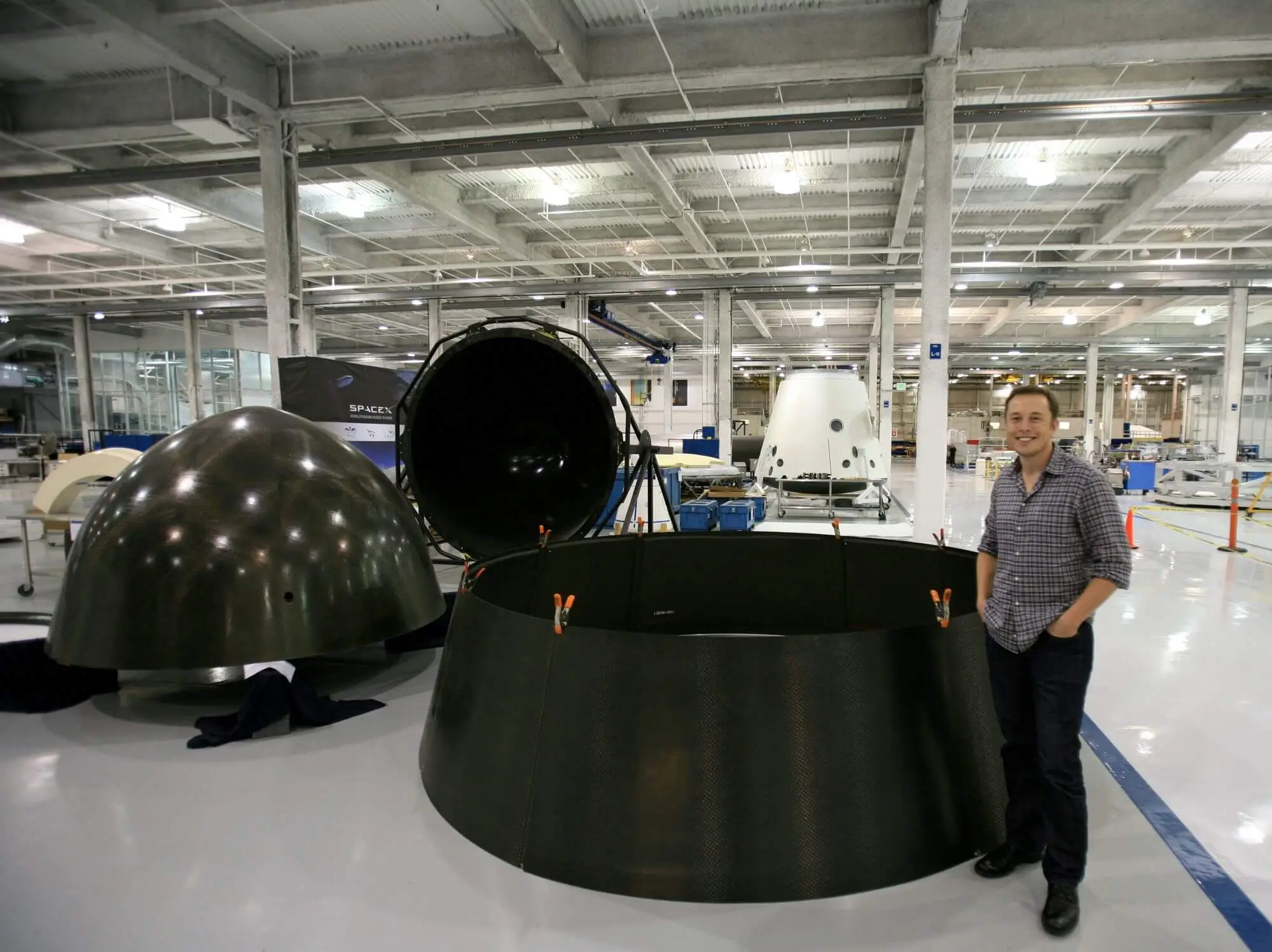 Elon Musk standing beside early SpaceX hardware, including a Dragon capsule and carbon-fibre components, in the company's factory